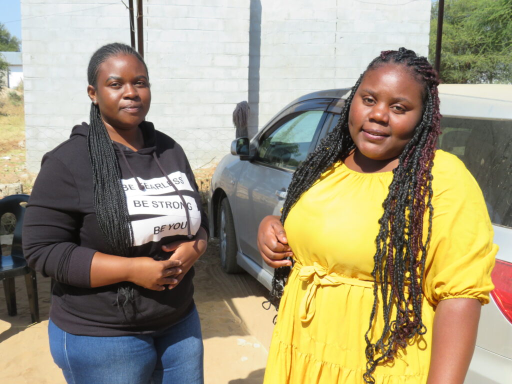 Two young women, one in a black hoodie that reads "be fearless, be strong be you" and the other in a vibrant yellow sundress, stand in a car park. They are project leaders at the Dreams Club in Divundu, which teaches young women and girls about family planning.
