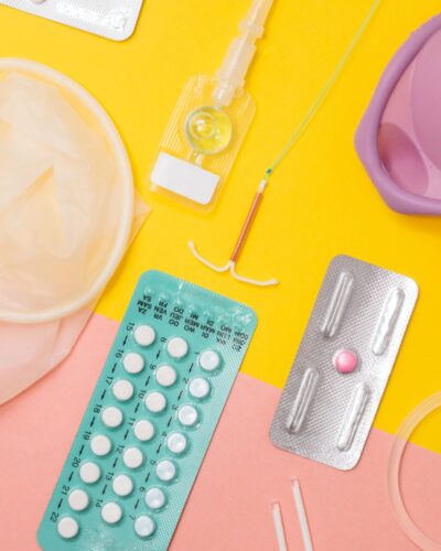A variety of contraception methods for people with the capacity for pregnancyare scattered on a background that is half sunny-yellow and half soft pink.