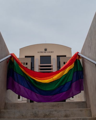 A Pride flag held aloft in the foreground, with the Namibian Supreme Court in the background.